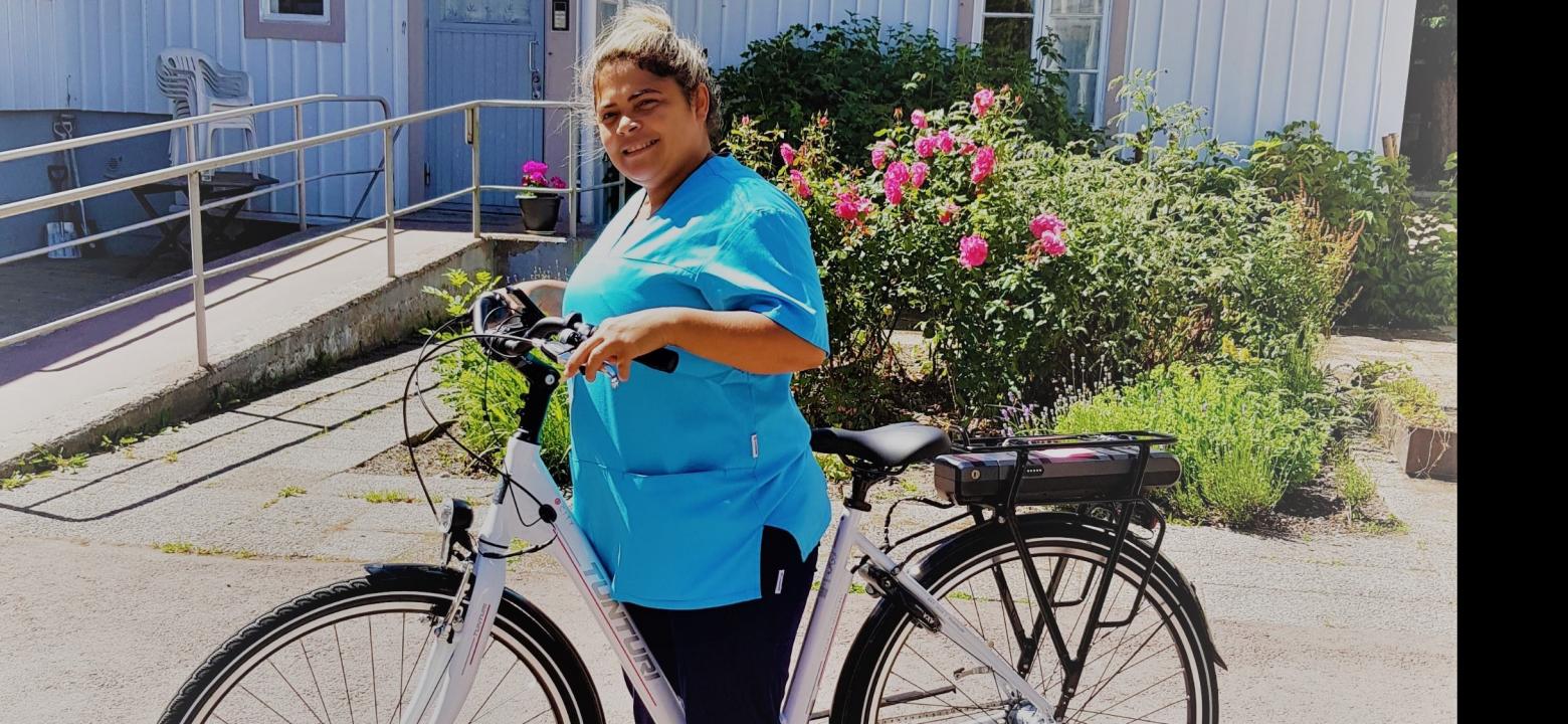 Närvårdare Maria med en utav de nya elcyklarna Närvårdare Maria i turkos arbetsdräkt står med en utav de nya vita elcyklarna, med en rosenbuske i bakgrunden.
