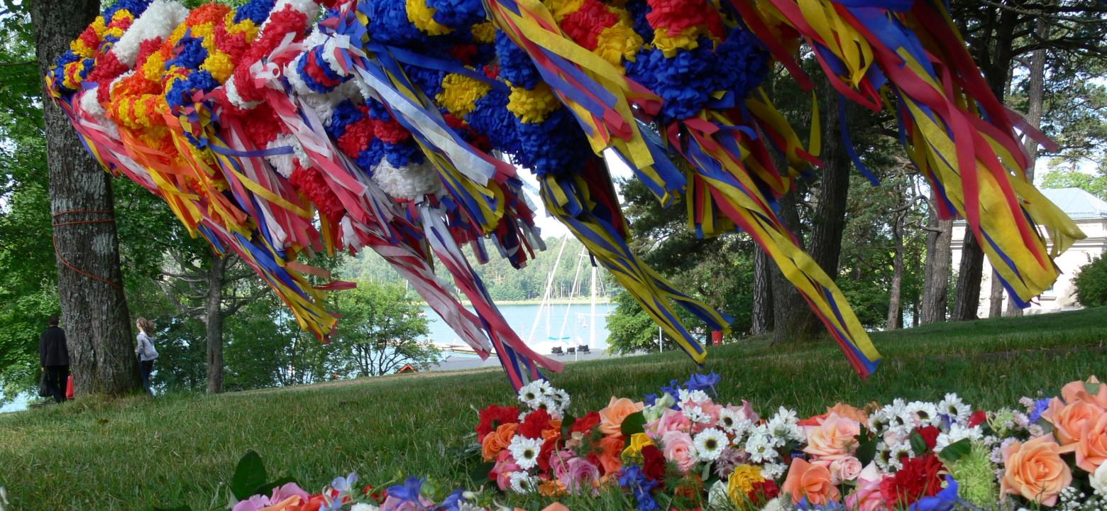 Foto på midsommarkronor och blomsterarrangemang i Engelska parken. I bakgrunden syns vatten och en segelbåt.