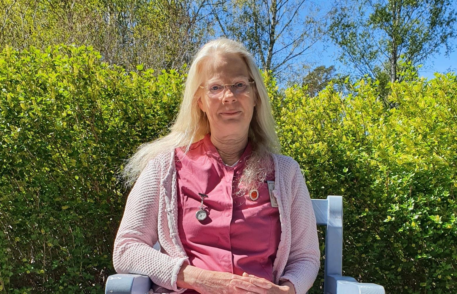 Bild på Mariehamns äldreomsorgs nyanställda Silviasyster Annica Rydberg, som sitter på en stol framför en grönskande häck. Hon har långt ljust hår och bär rosa arbetstunika med stickad tröja.