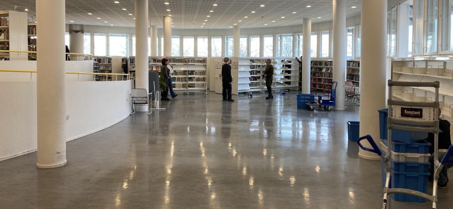 Foto på bibliotekets skönavdelning tom på hyllor och böcker. Folk som arbetar med att flytta bokhyllor. 