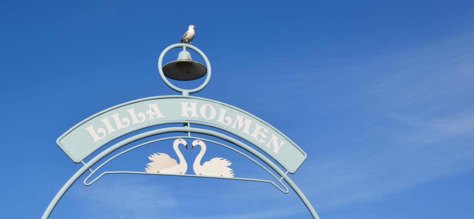Skylt vid bron till Lilla holmen med en klocka överst och två svanar under texten Lilla holmen