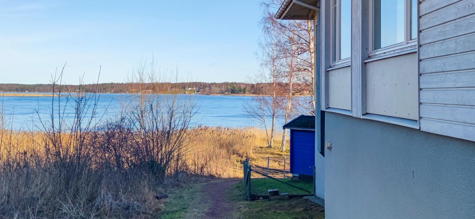 Bilden visar att Strandbacken har stranden precis nedanför backen, här visas bara hörnet av huset och att gården är inhägnad och en blå lekstuga syns.