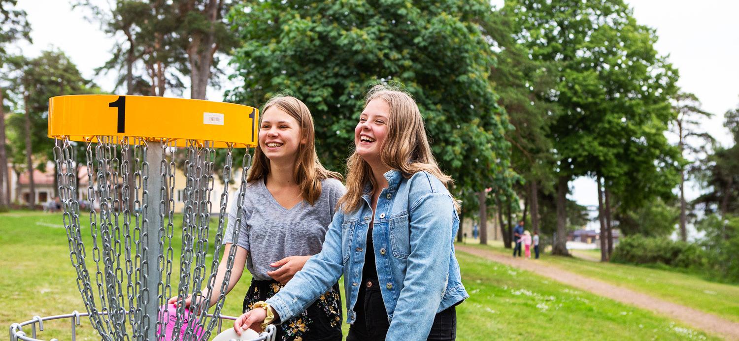 Bilden visar två skrattande tonårstjejer som spelar discgolf i Badhusparken