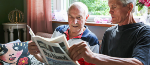 Bilden visar en anställd vid hemservice som läser en tidning för en äldre man i hans hem