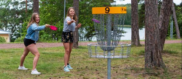 Bilden visar två unga tjejer spelandes discgolf bland träd i en park med havet i bakgrunden